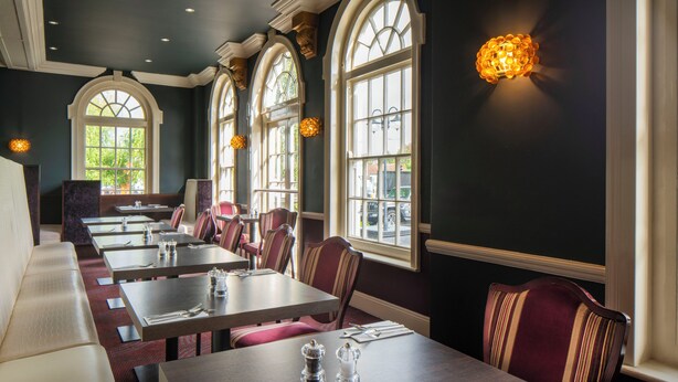 En restaurang med många bord och avskilda sittplatser, stora fönster och dekorativa vägglampor.