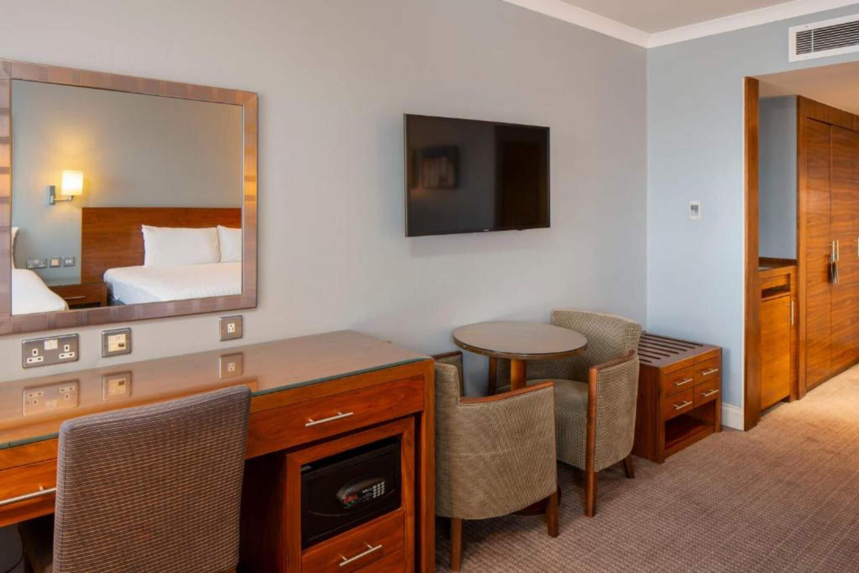 Une chambre d’hôtel comprenant un lit, un bureau avec un miroir, une télévision, une petite table ronde avec des chaises et une armoire.