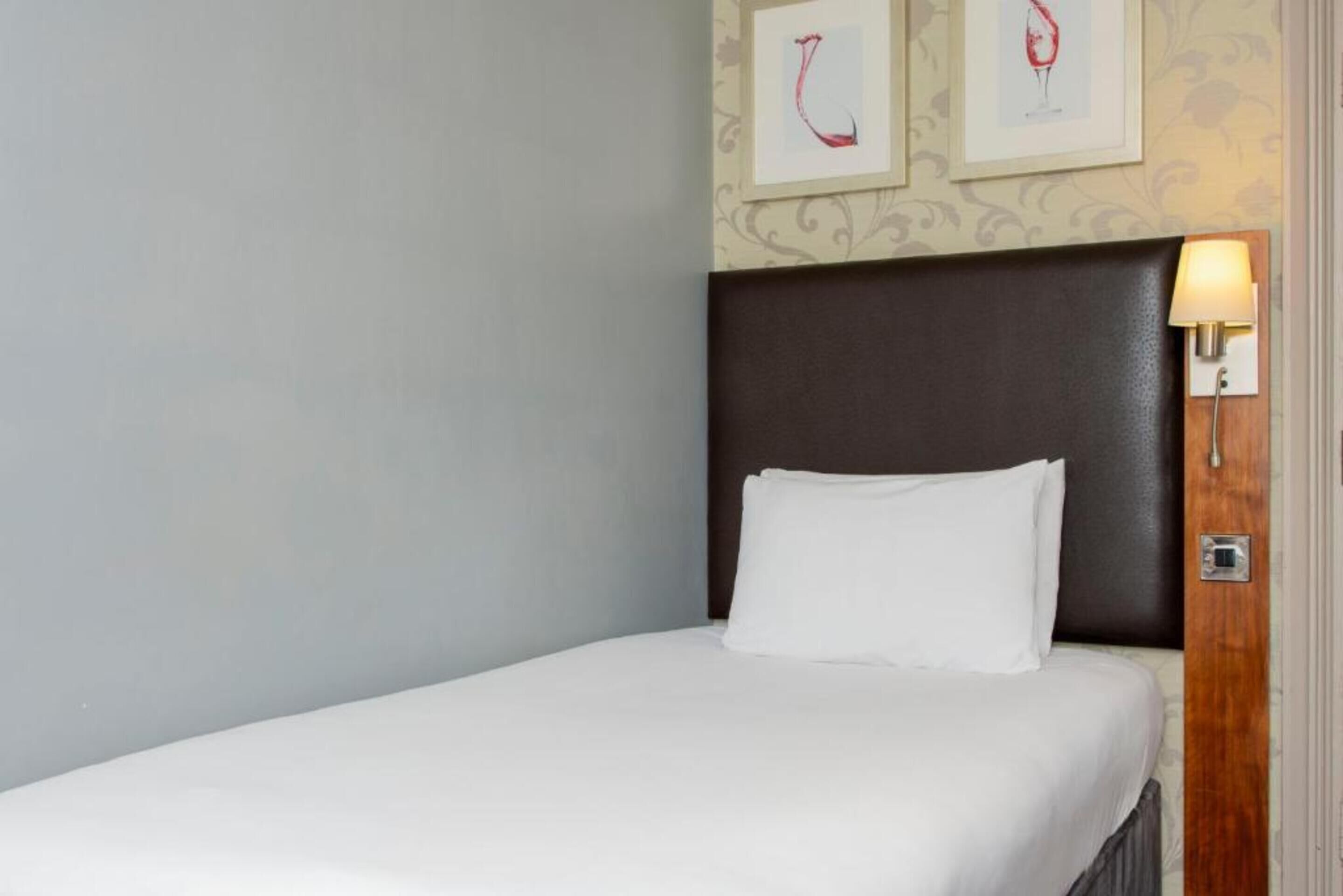 A hotel room with a single bed, a dark headboard, and a wall-mounted light fixture.