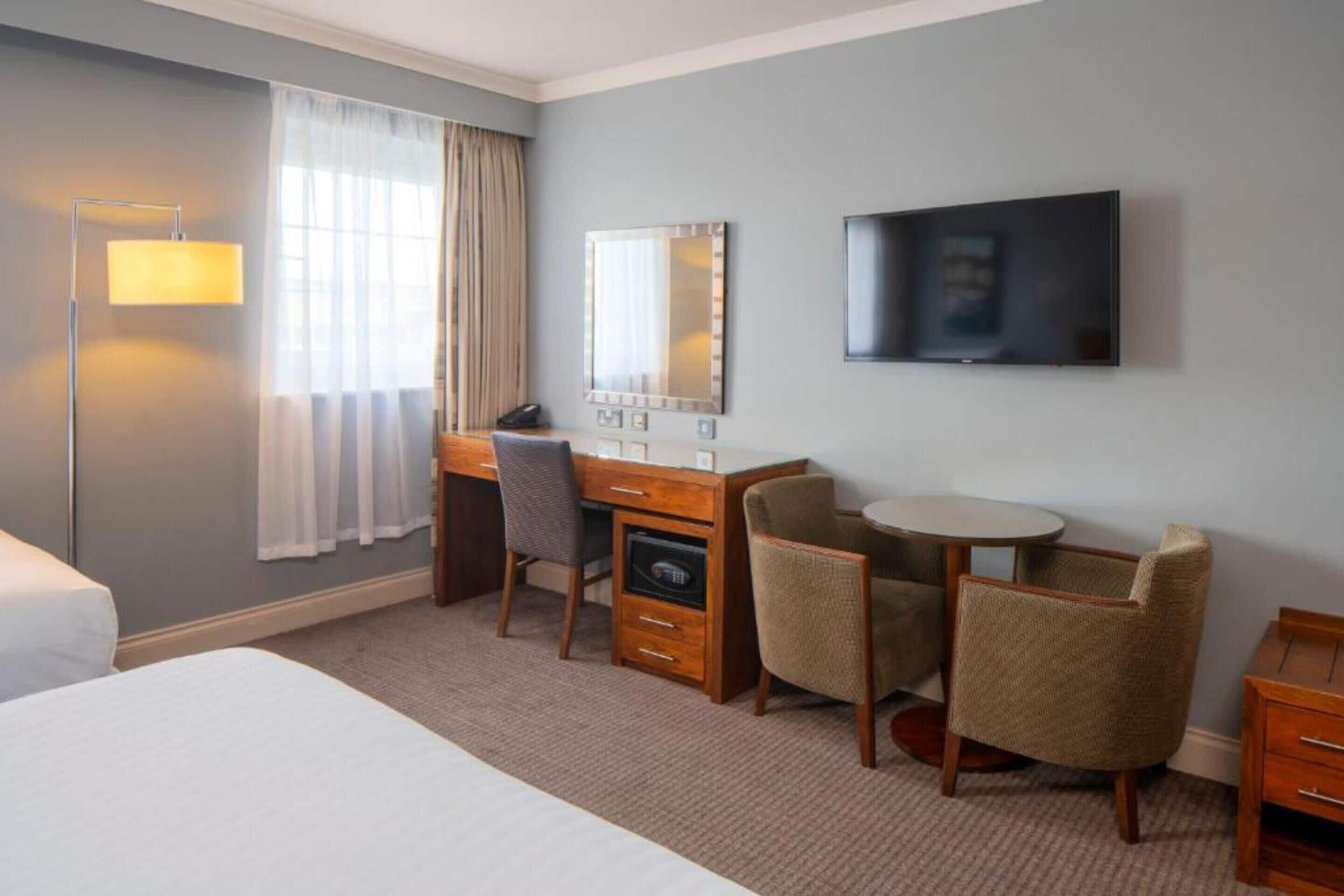 A hotel room with a bed, a desk with a chair, a round table, a TV, and a mirror.