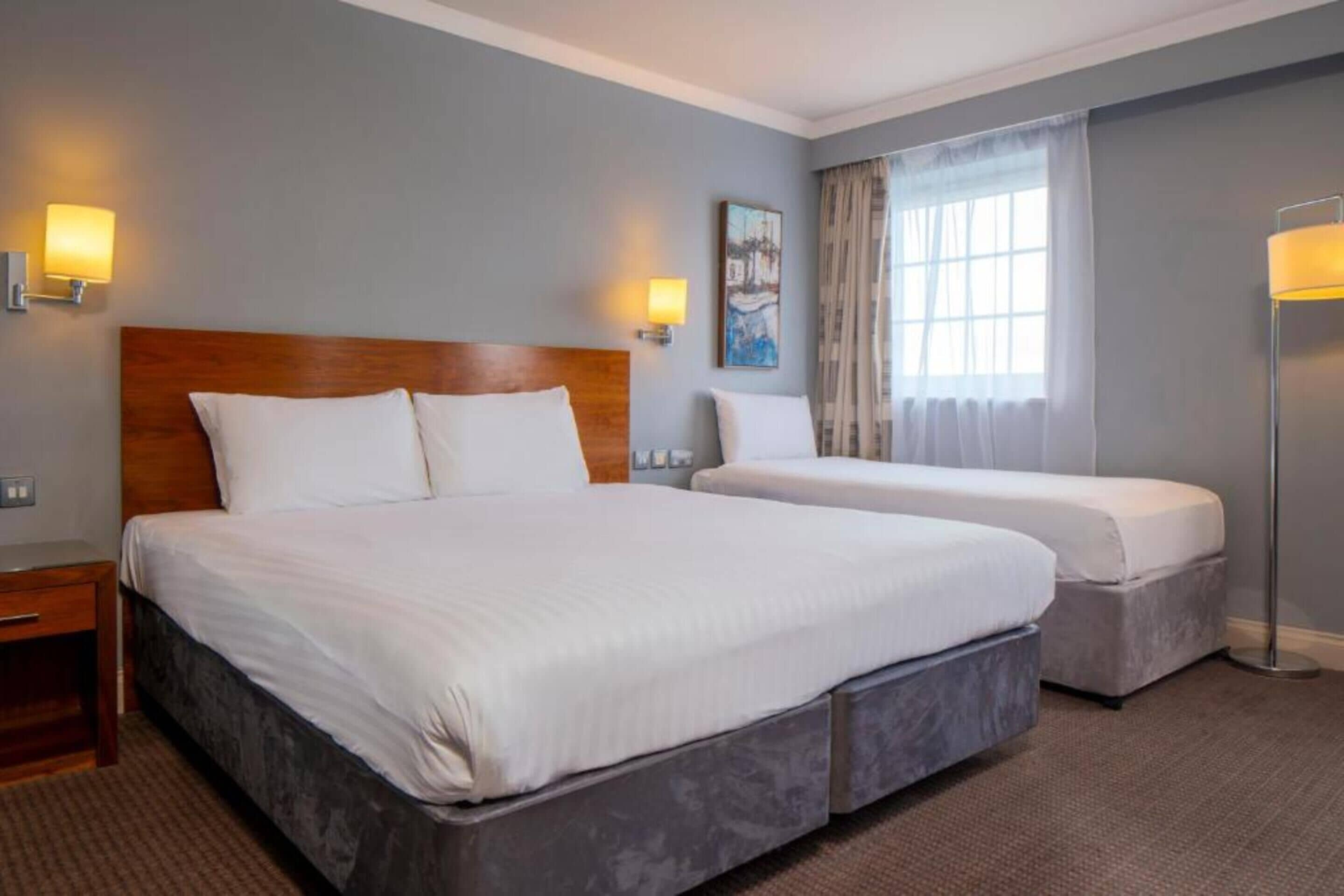 A hotel room with a large bed, a smaller bed, a nightstand, a lamp, and a painting on the wall.