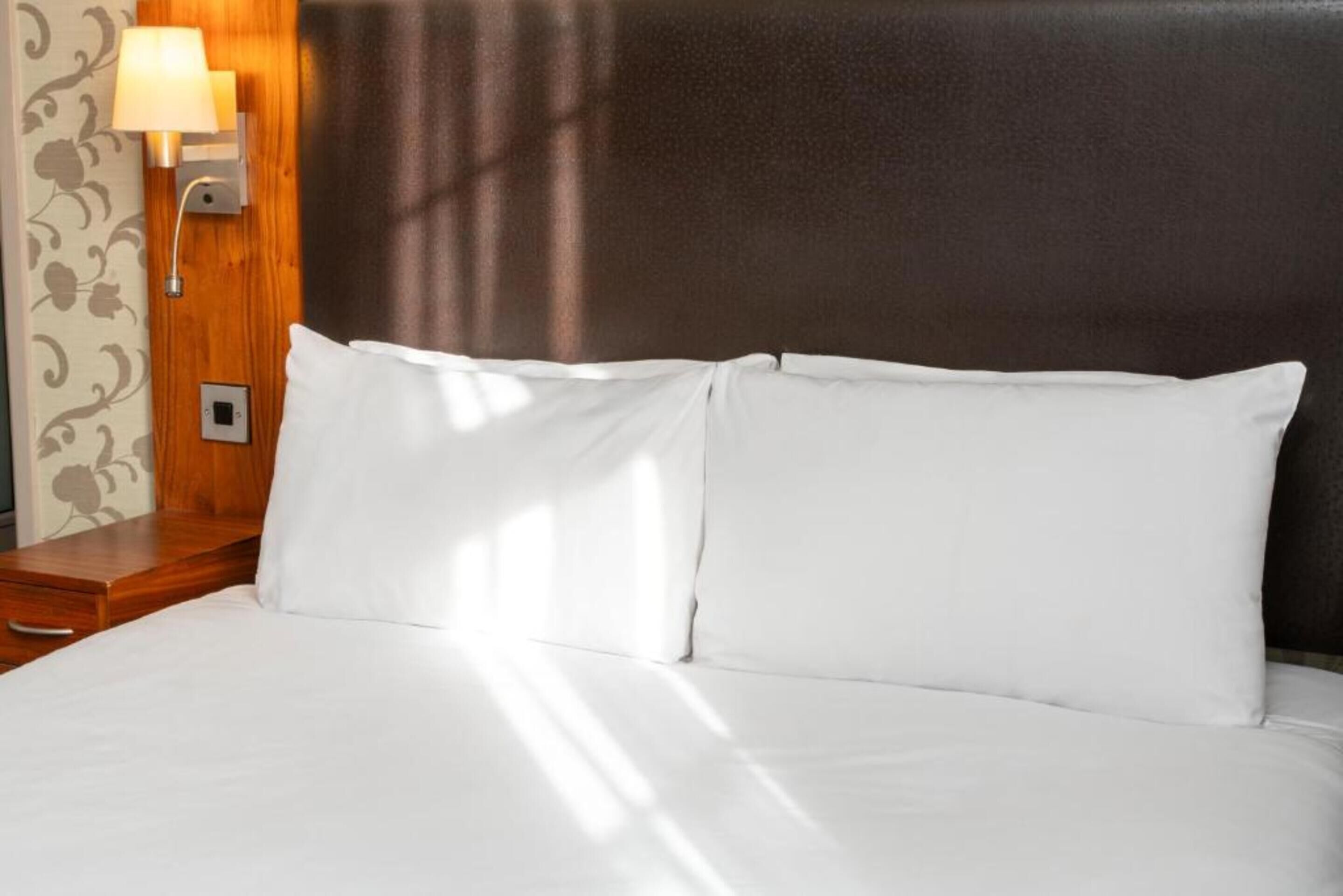 A neatly made bed with white linens, a wooden nightstand with a lamp, and a wall with floral wallpaper.