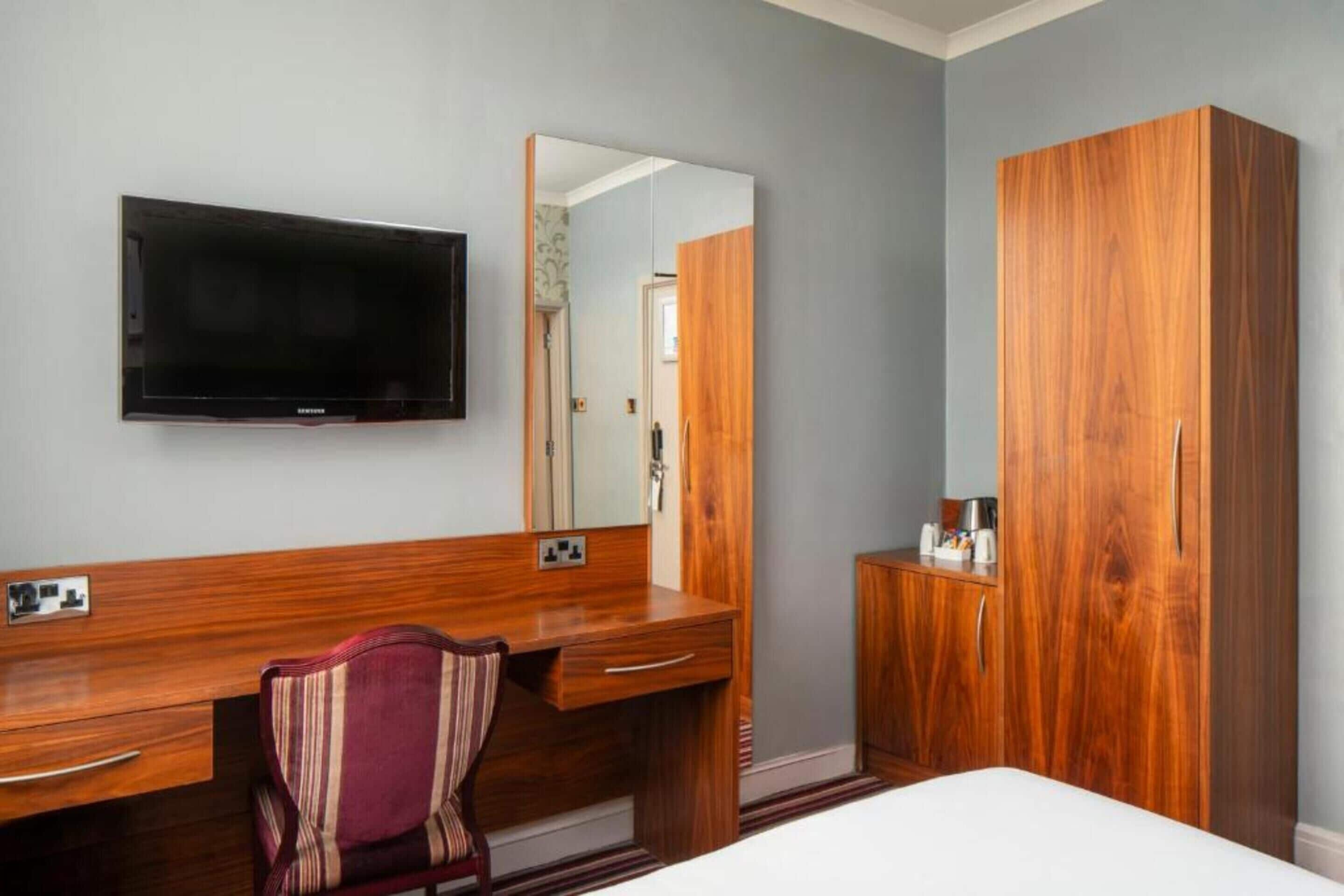 Ein Hotelzimmer mit Holztisch, Wandfernseher, großem Spiegel, Kleiderschrank und Bett.