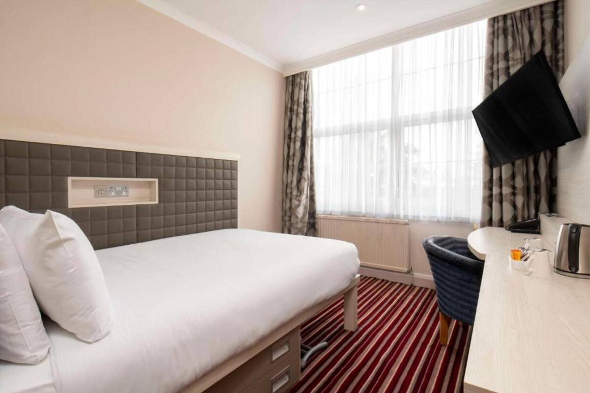 Camera d'albergo con un letto, una scrivania, una sedia, una TV a parete e una finestra con tende.