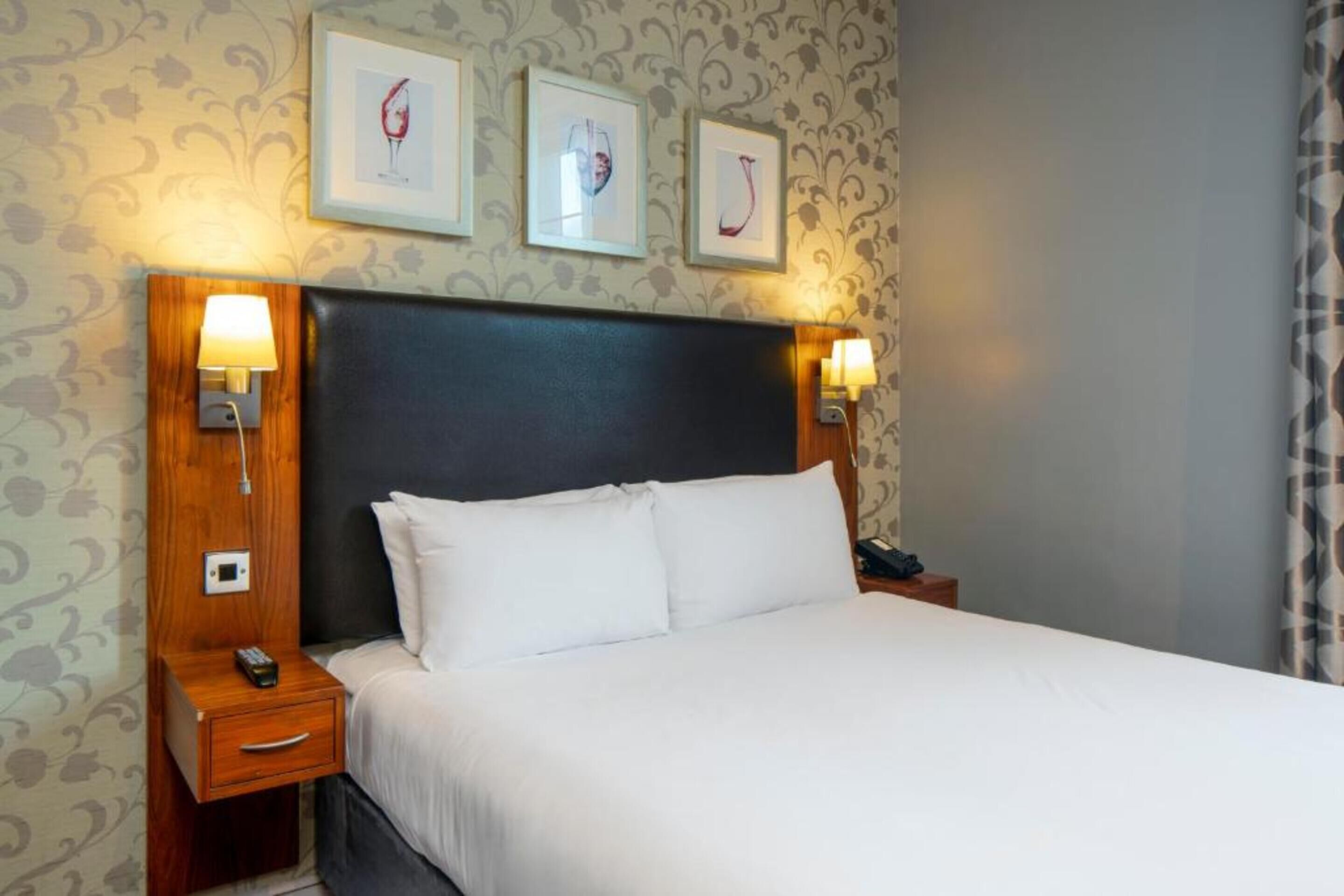 Une chambre d’hôtel comprenant un lit, deux tables de chevet avec des lampes, une petite table et trois tableaux encadrés accrochés au mur.