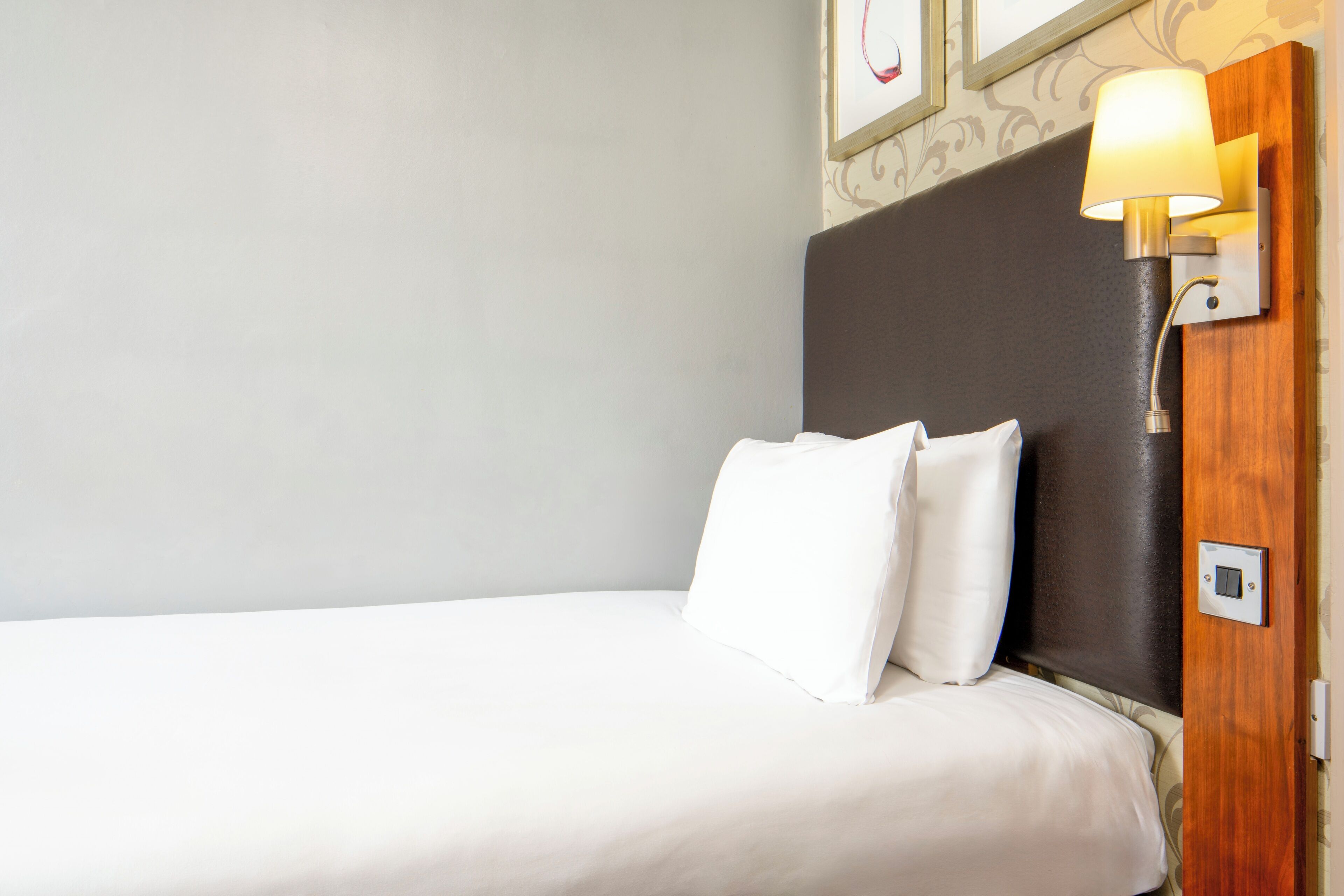 A neatly made bed with white linens and pillows, a dark headboard, and a wall-mounted lamp.