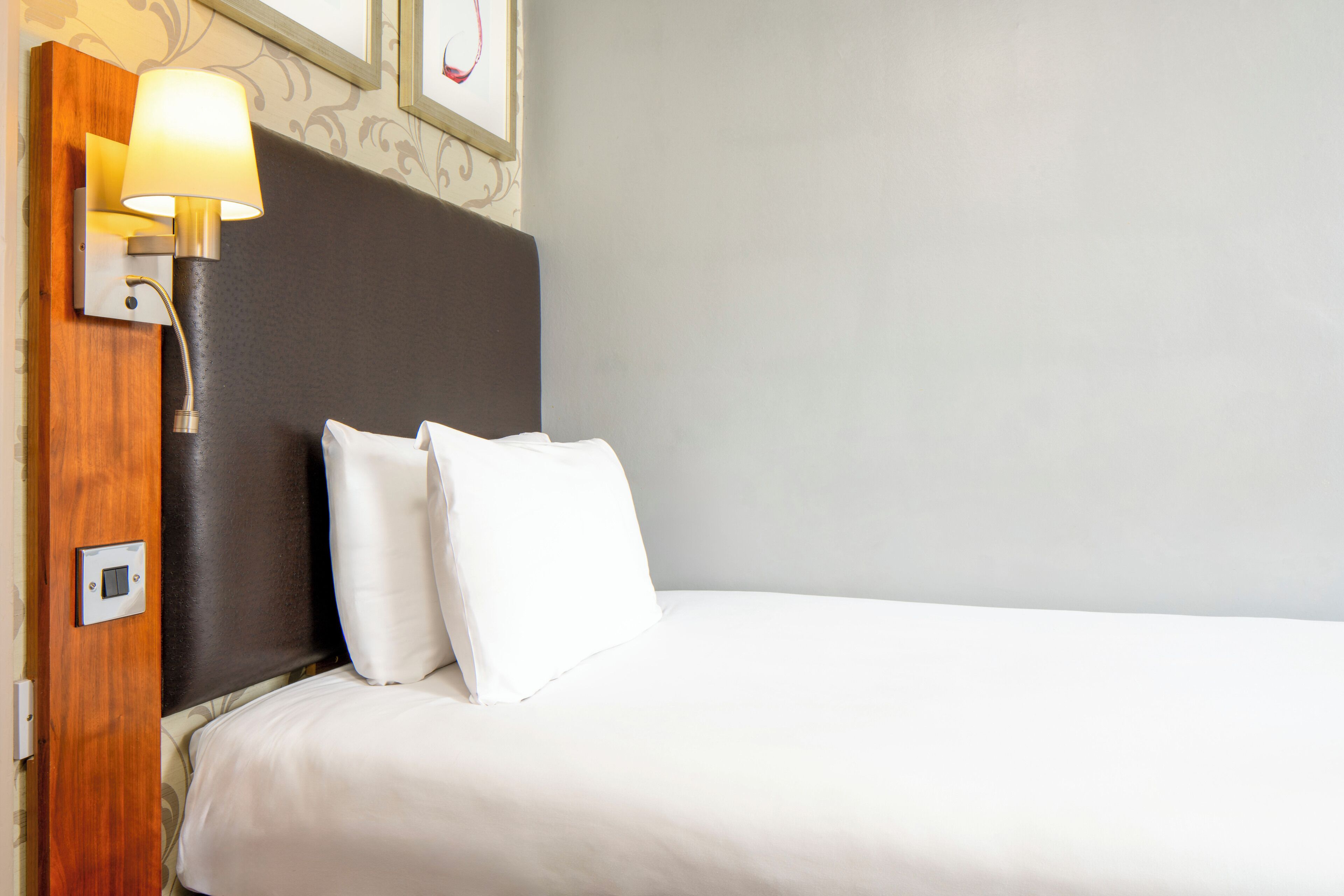 Une chambre d’hôtel avec un lit, du linge de lit blanc, une tête de lit sombre, une lampe fixée au mur et un tableau encadré au-dessus du lit.