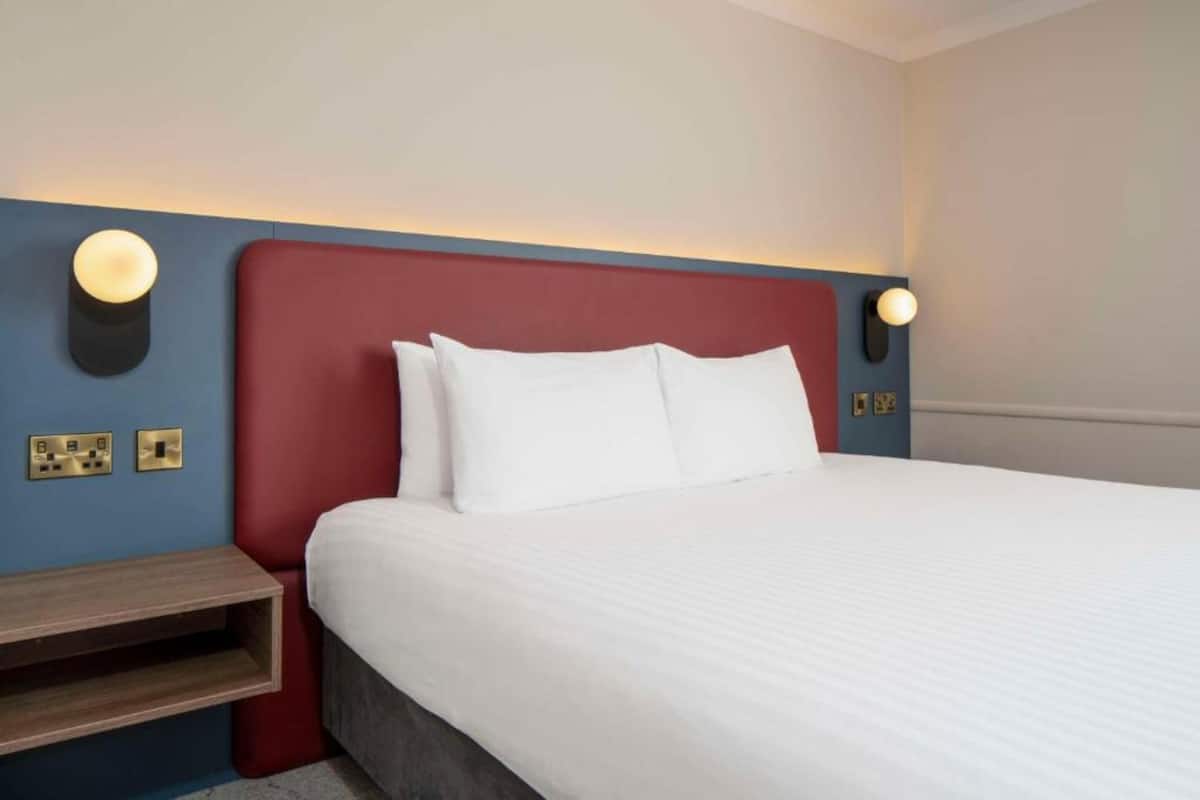 Una camera d'albergo con un letto, un comodino, una lampada a parete e prese elettriche.