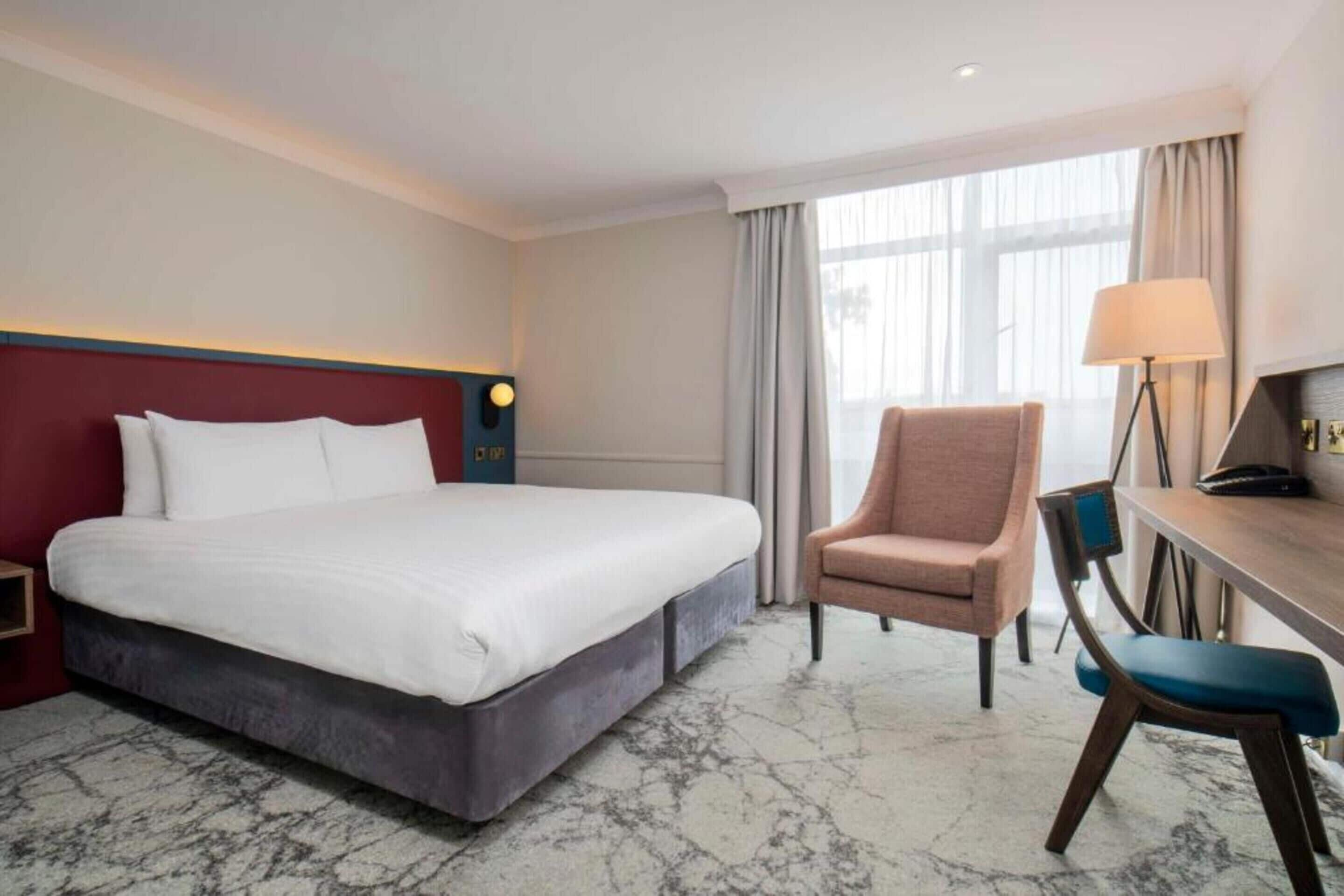 A hotel room with a large bed, a desk with a chair, a lamp, and a window with curtains.