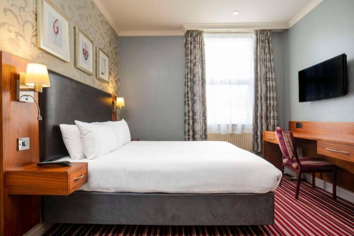 Una camera d'albergo con un letto, comodini, una scrivania, una sedia, un televisore e quadri appesi al muro.
