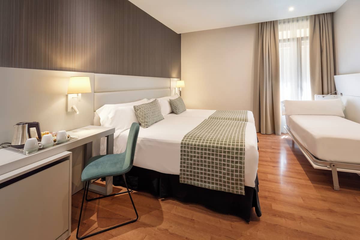 A hotel room with a bed, a desk with a chair, a coffee maker, and a window with curtains.