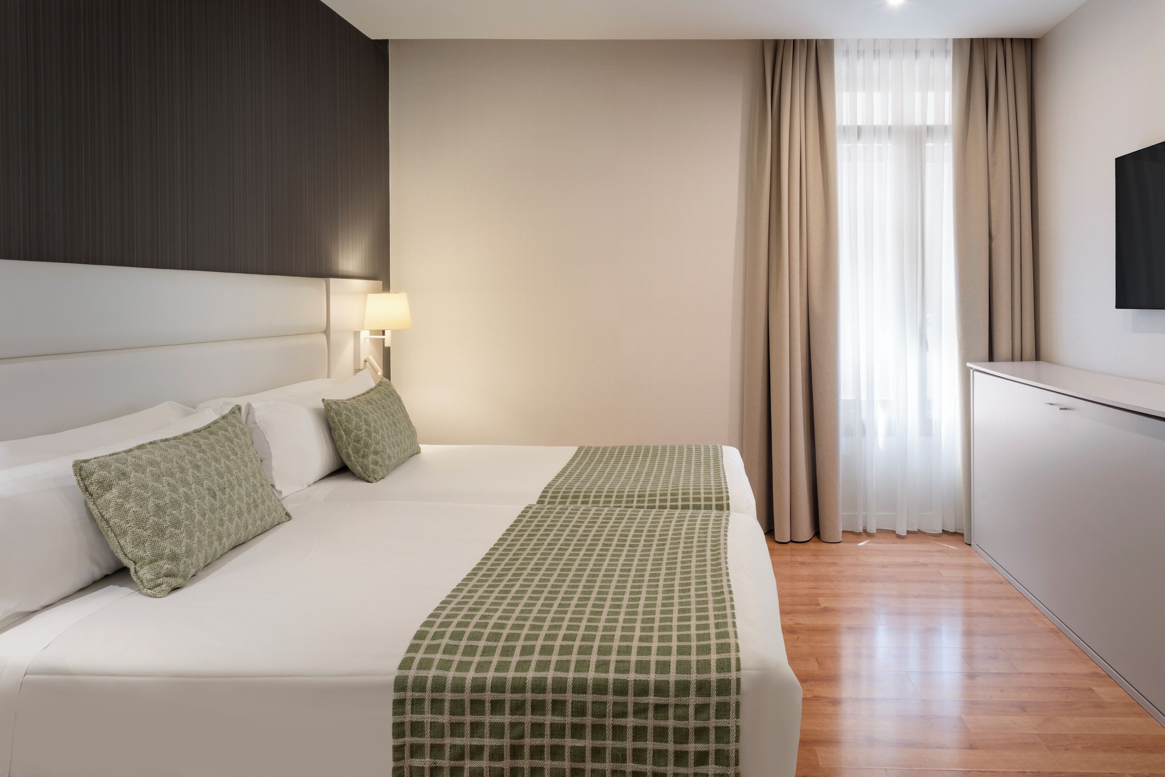 A hotel room with a large bed, a bedside table with a lamp, a TV mounted on the wall, and a window with curtains.
