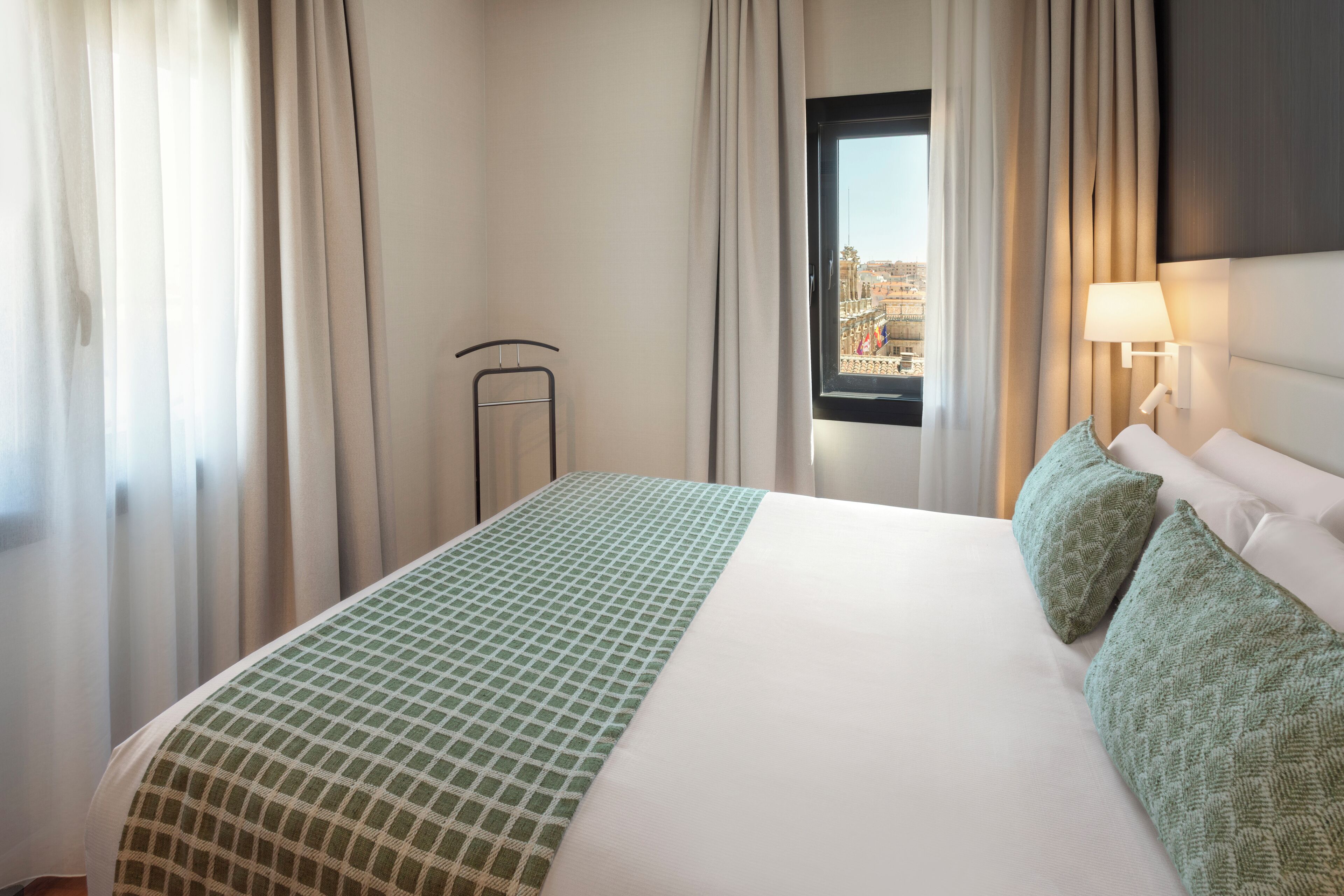 Una habitación de hotel con una cama grande, una ventana con cortinas, una lámpara y vistas a un paisaje urbano.