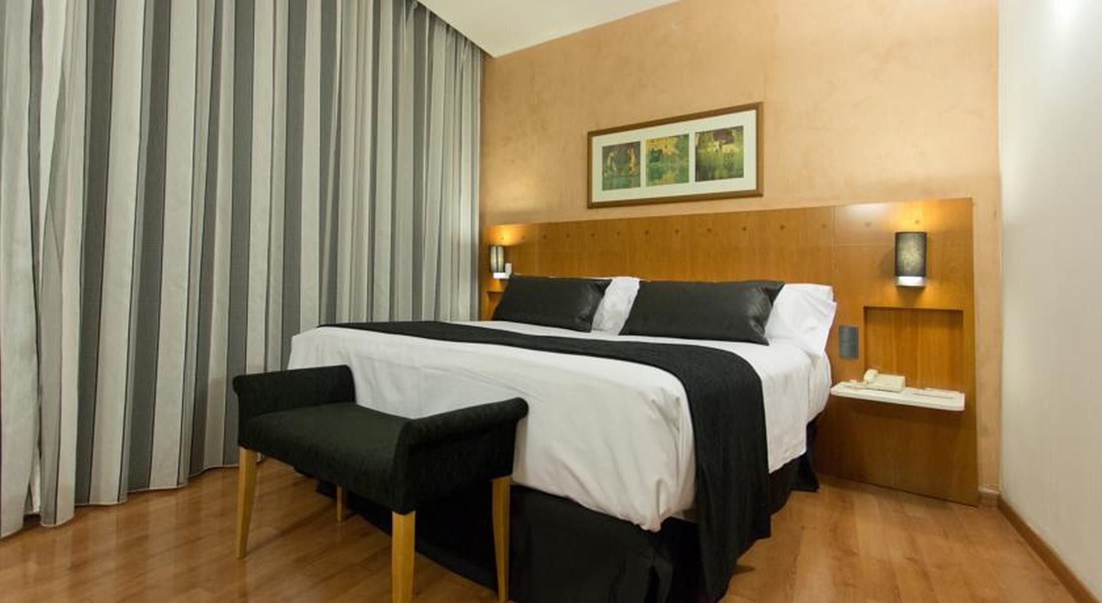 Une chambre d’hôtel avec un lit, une banquette, une table de chevet et un tableau accroché au mur.