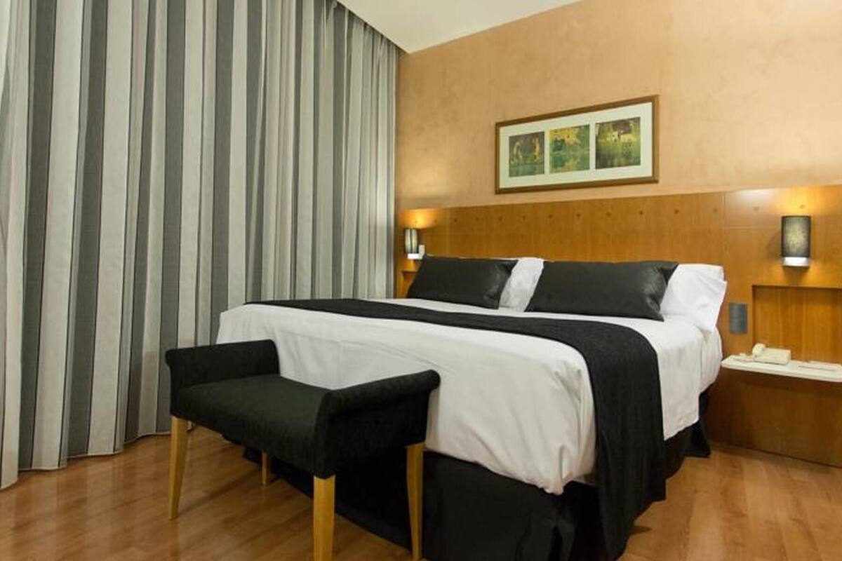 Une chambre d’hôtel avec un lit, une banquette, une table de chevet et un tableau accroché au mur.