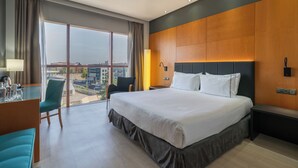 Quarto de hotel moderno com uma cama grande, uma escrivaninha com cadeira, vista para a cidade e uma janela ampla com cortinas.