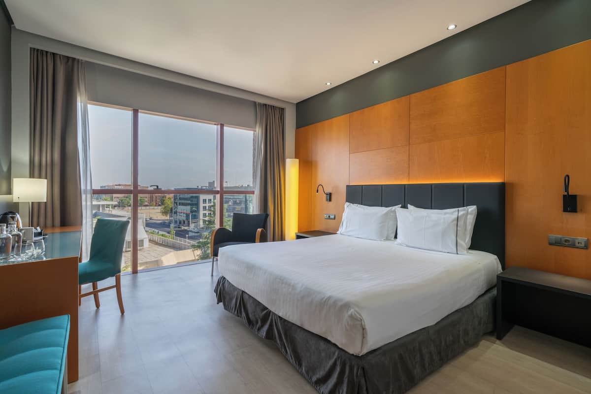A modern hotel room with a large bed, a desk with a chair, a view of the city, and a large window with curtains.