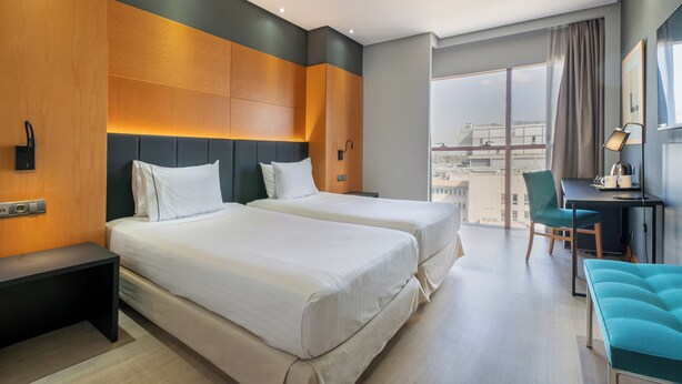 Quarto de hotel com duas camas, uma escrivaninha, uma cadeira e vista para a cidade.