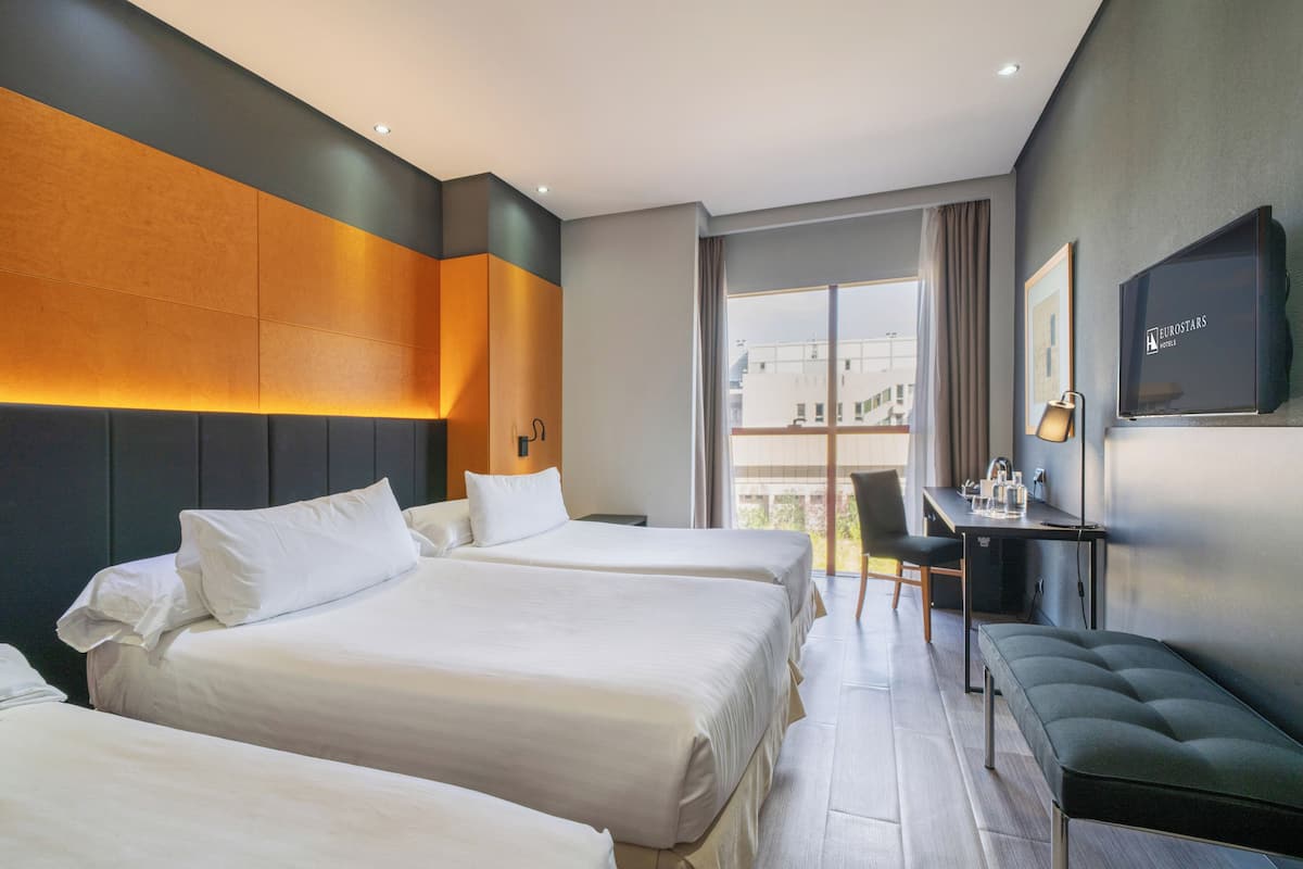 A hotel room with three beds, a desk, a bench, a TV, and a large window.