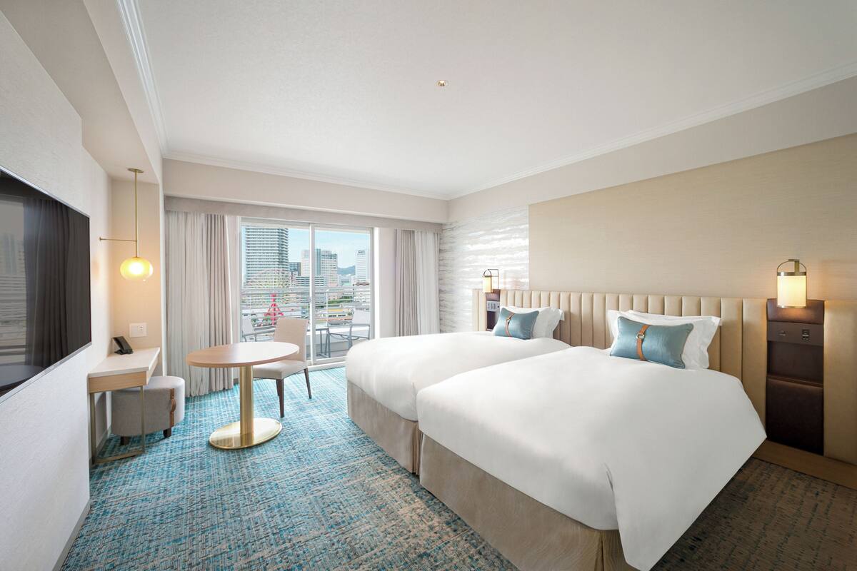 Habitación de hotel con dos camas, un escritorio, una silla, una mesa redonda y vistas de la ciudad a través de amplias ventanas.