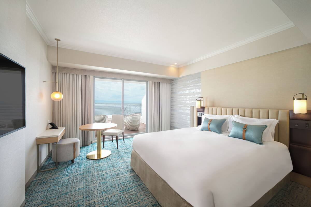 Habitación de hotel con una cama grande, un escritorio, una silla, una mesita y vista al océano por la ventana.