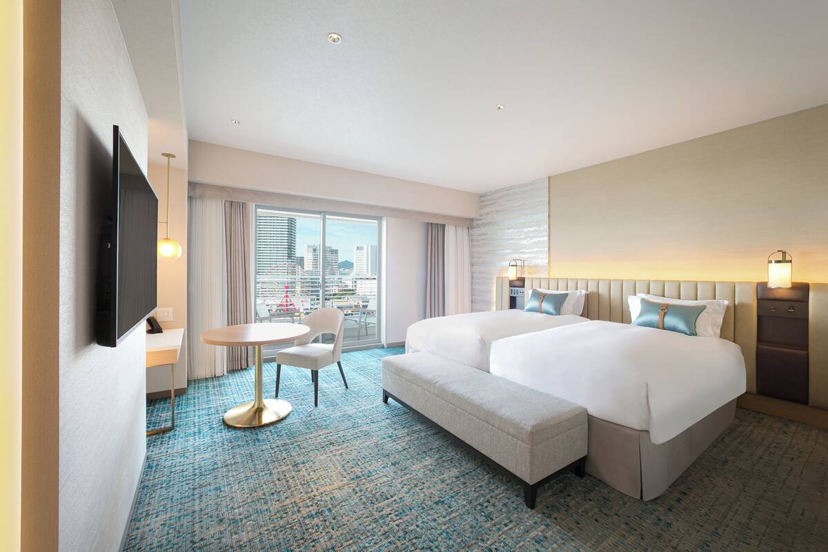 Habitación de hotel con una cama grande, un escritorio con una silla, un televisor y vista a la ciudad por la ventana.