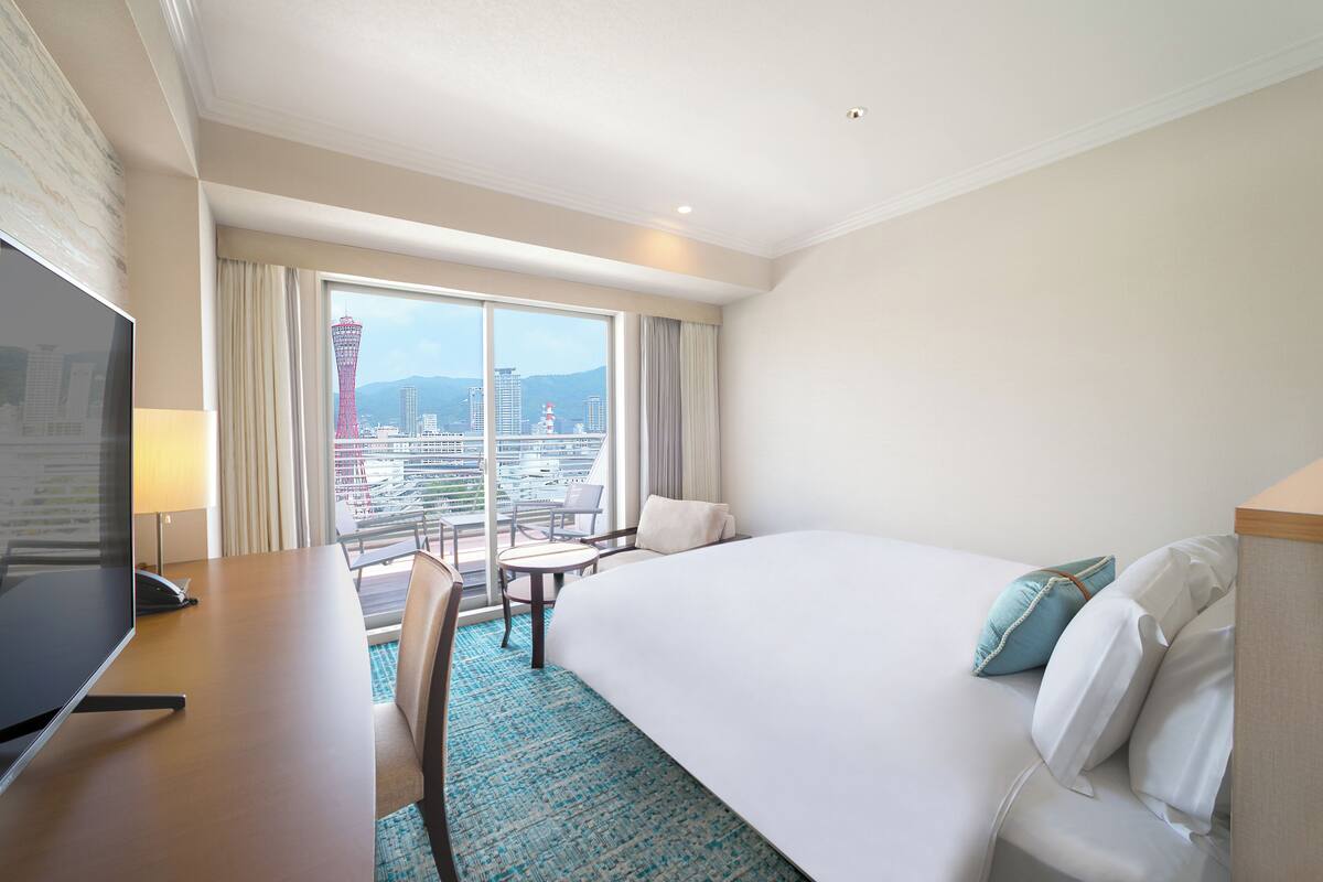 Habitación de hotel con una cama grande, un escritorio con televisor y vistas a la ciudad a través de amplias ventanas.