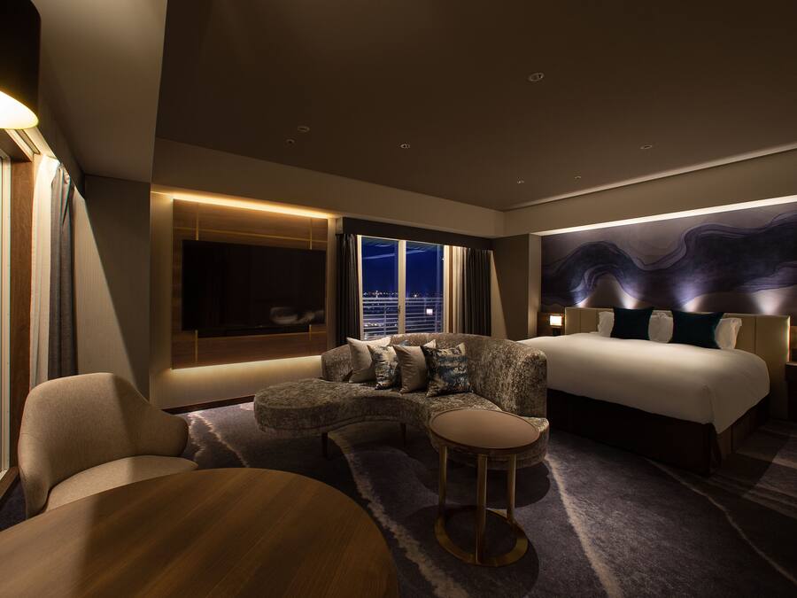 Quarto de hotel moderno com sofá curvo, mesa redonda, cama e vista da cidade à noite.