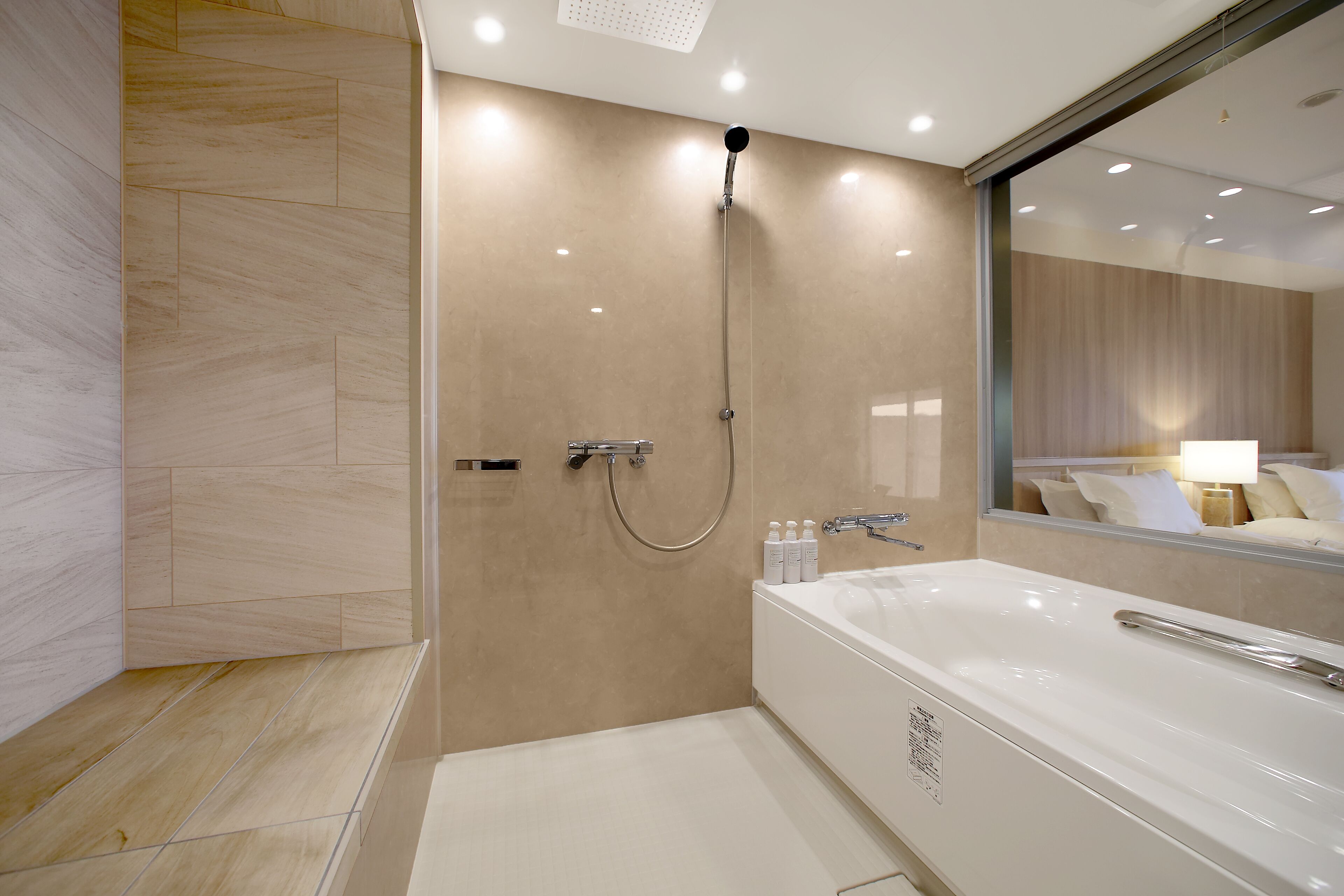 A modern bathroom with a walk-in shower, a large bathtub, and a mirror reflecting a bed with pillows.