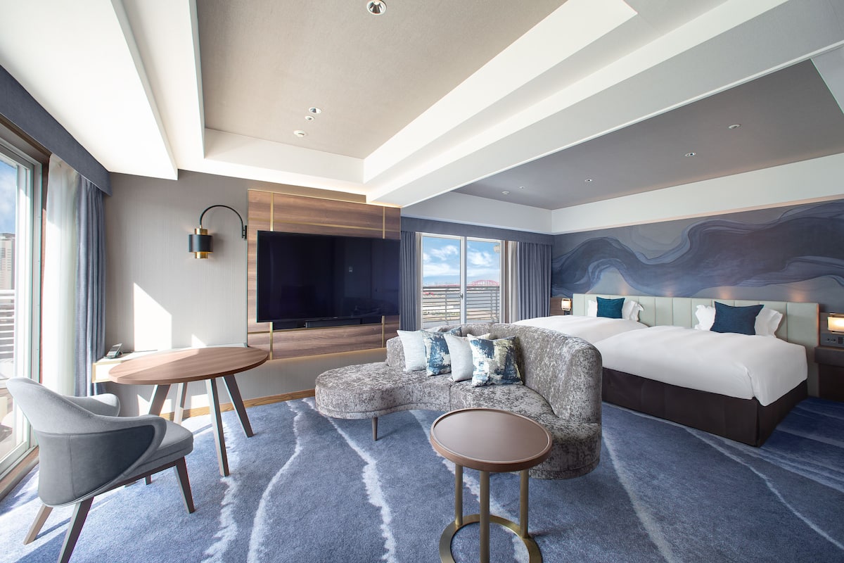 Una habitación de hotel moderna con un sofá curvo, una mesa de centro redonda, una cama con cabecera y un ventanal con vistas a la ciudad.