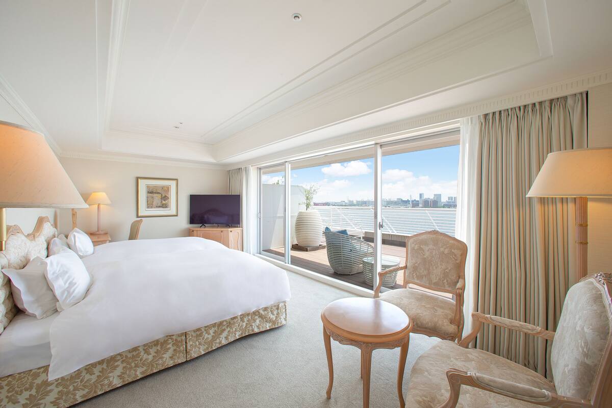Habitación de hotel con una cama grande, dos sillones, una mesita, un televisor y un balcón con vistas a la ciudad.