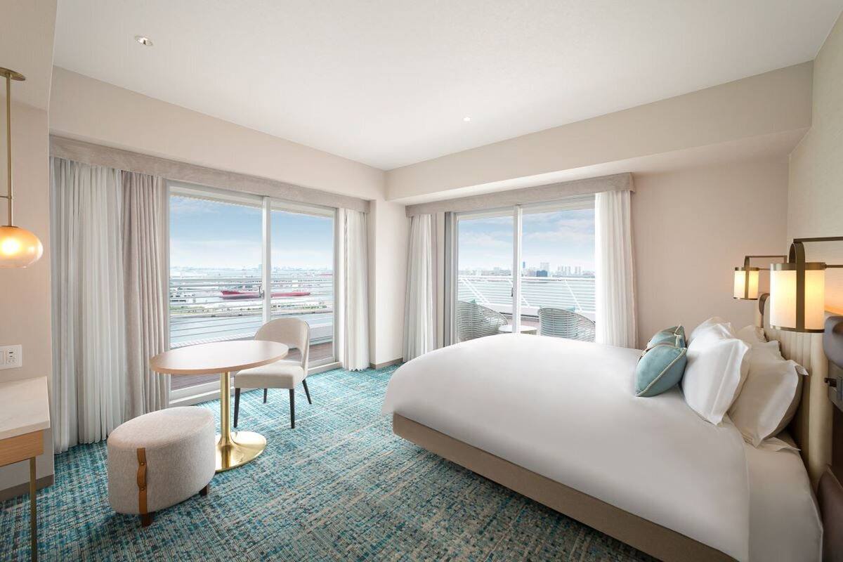 Habitación de hotel con una cama grande, una mesa redonda y dos sillas. Amplias ventanas ofrecen vistas a un paisaje urbano y un puente.