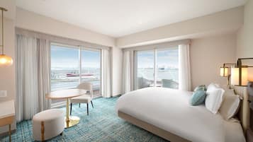 A hotel room with a large bed, a round table, and two chairs. There are large windows with a view of a cityscape and a bridge.