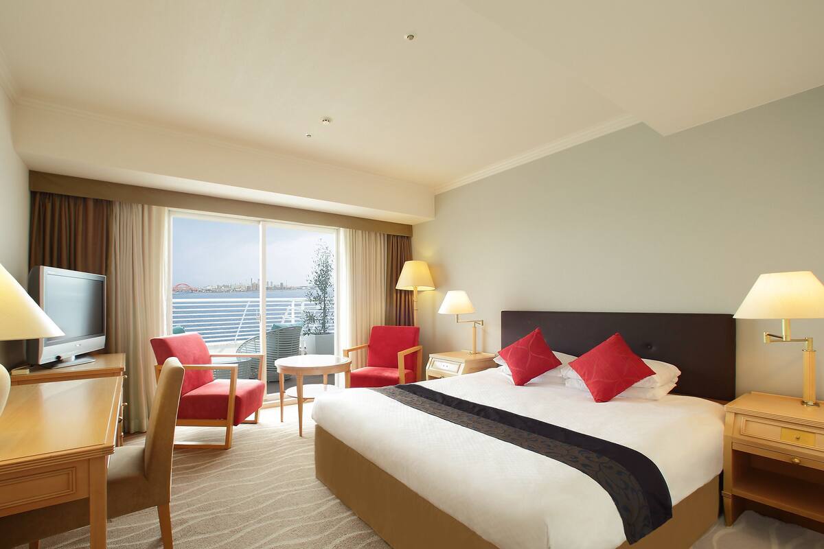 Habitación de hotel con una cama grande, dos sillas rojas, una mesita, televisión y vistas a un puente y al agua.