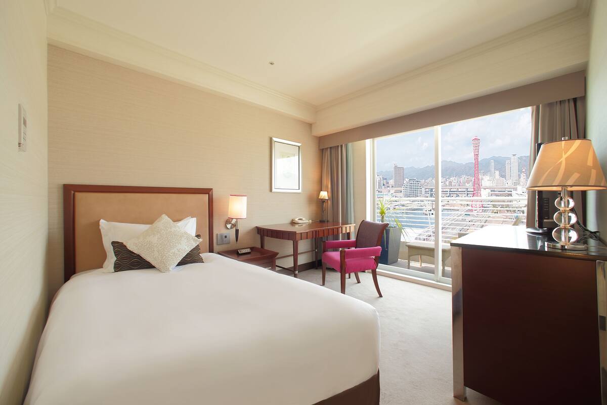 Habitación de hotel con una cama grande, un escritorio, una silla y vistas a un paisaje urbano a través de la ventana.