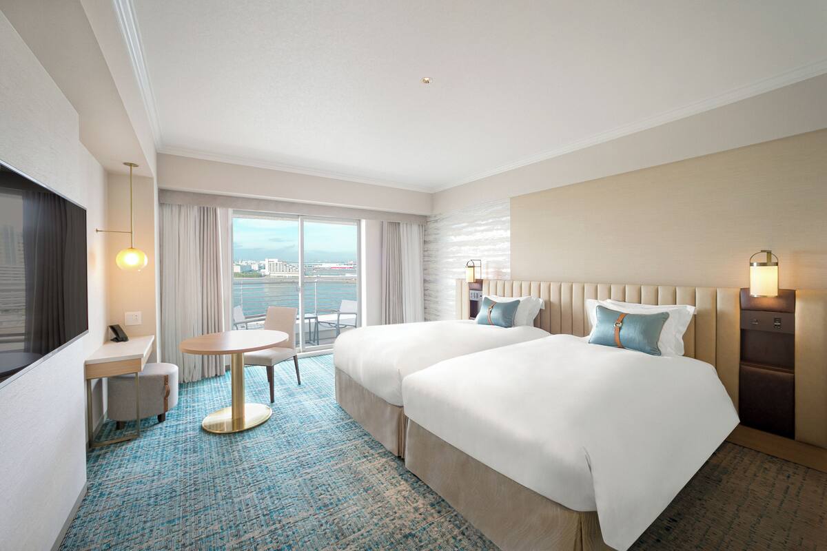 Habitación de hotel con dos camas, un escritorio, una silla, una mesita y un ventanal grande con vistas a la ciudad.