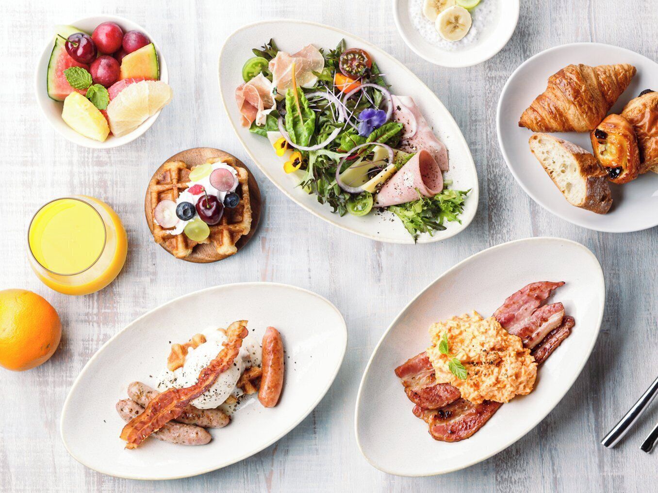 A breakfast spread with various dishes including a fruit bowl, a salad, waffles, croissants, and bacon.