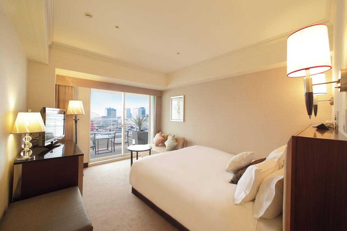 Habitación de hotel con una cama grande, un escritorio con lámpara, una silla, una mesita y vistas a la ciudad a través de un ventanal amplio.