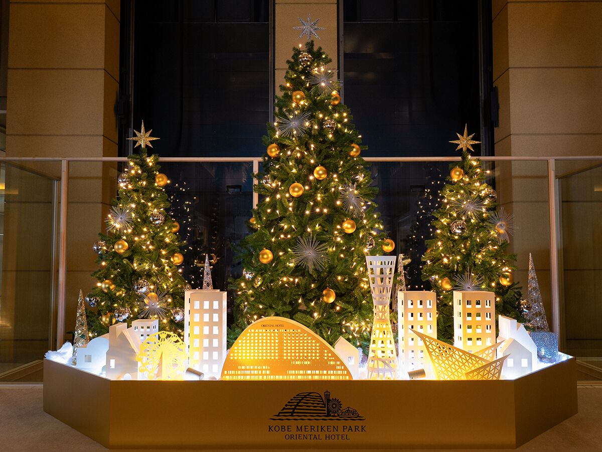 Une exposition de trois sapins de Noël décorés de lumières et d’ornements, le tout mis en valeur par la façade d’un bâtiment en arrière-plan.