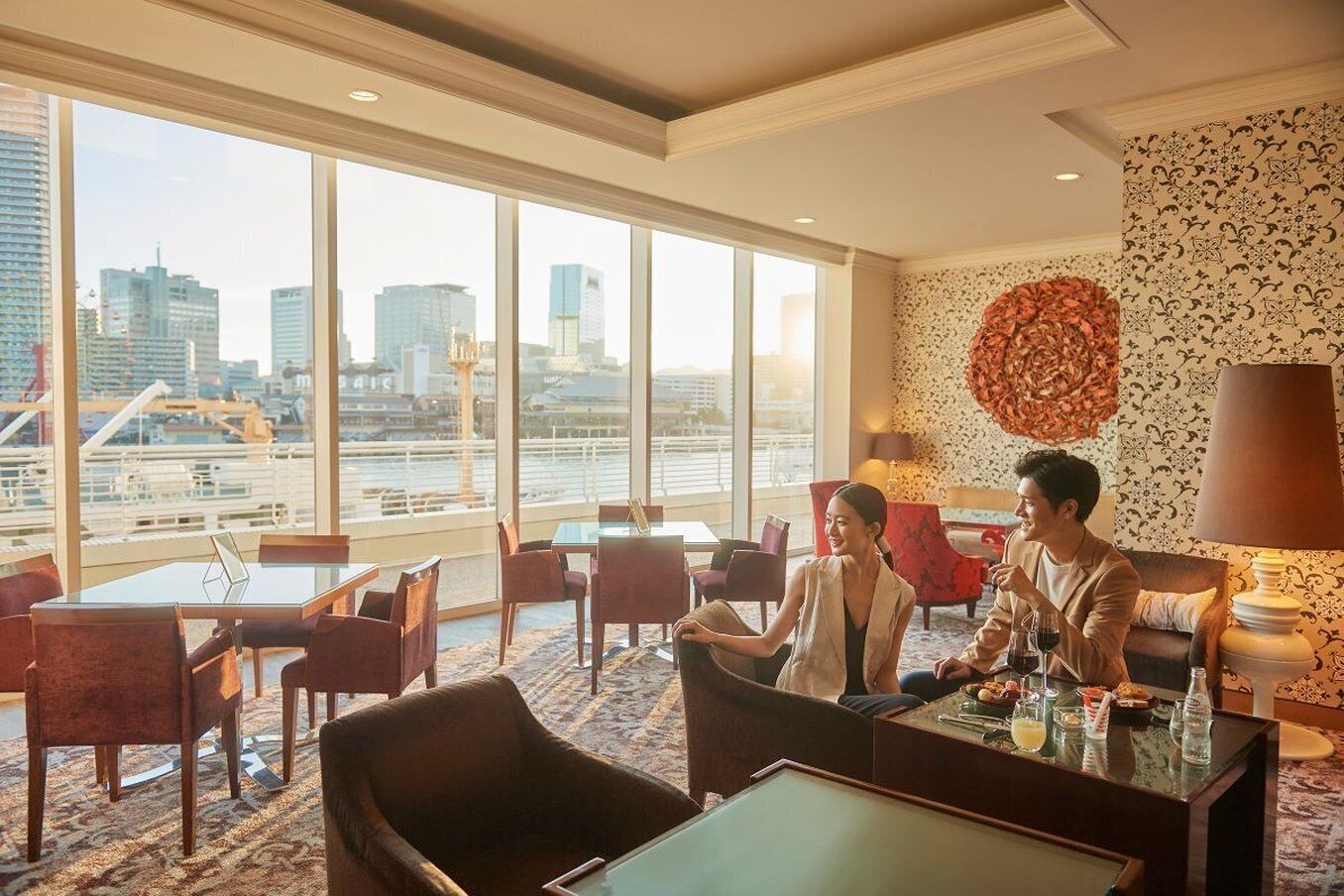 A modern hotel lobby with large windows offering a city view, comfortable seating, and a decorative wall piece.
