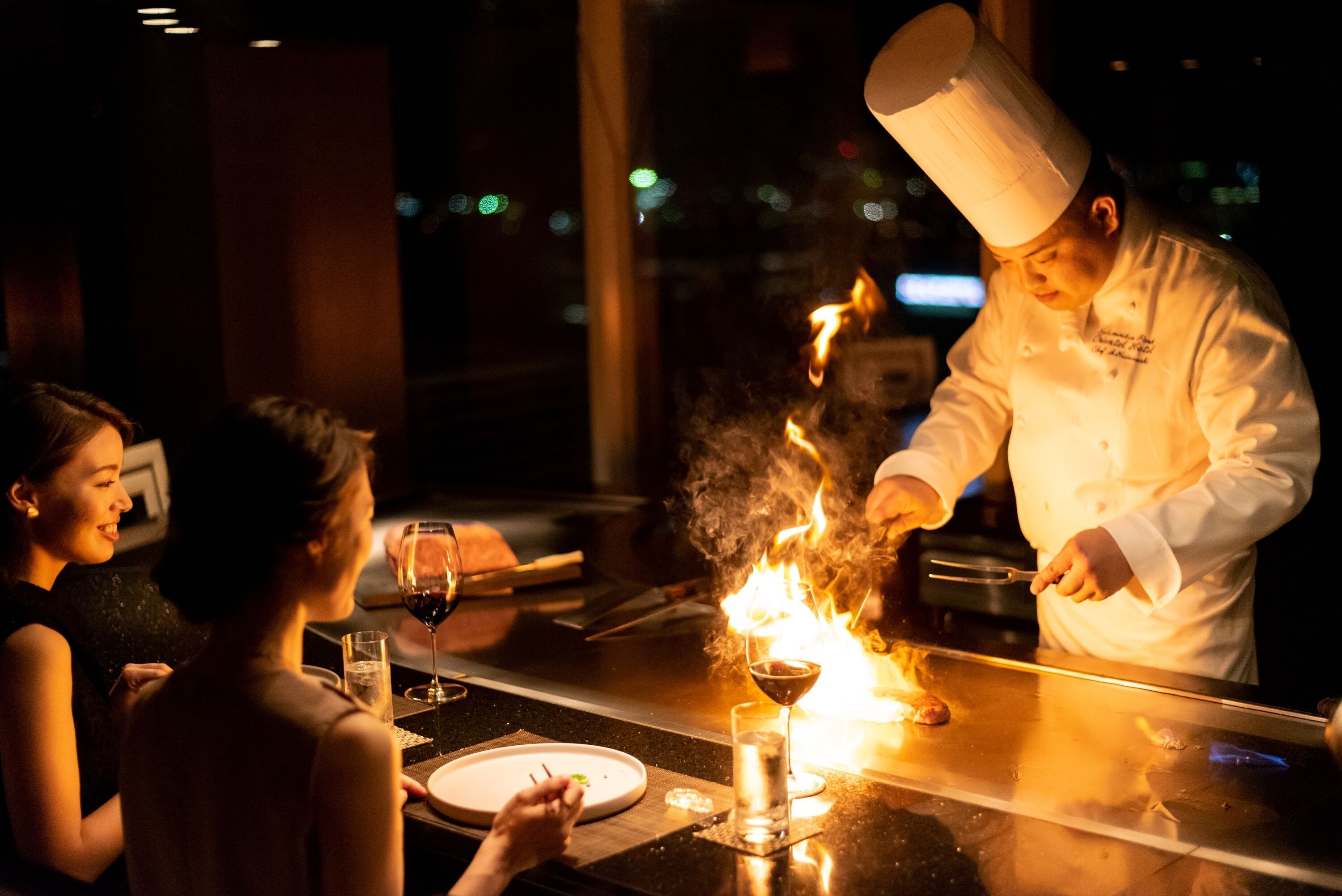 A chef performing a flambé at a restaurant table with diners watching.