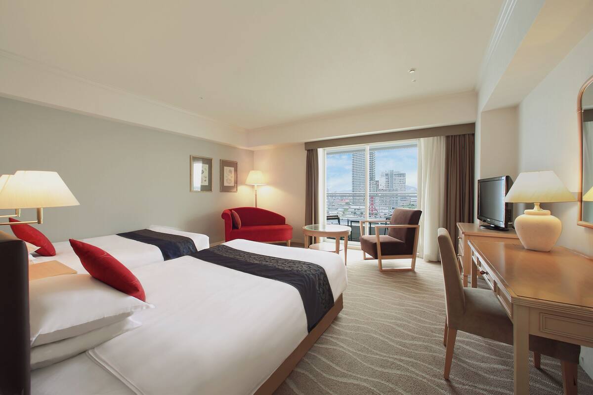 Habitación de hotel con dos camas, un escritorio, una silla, un televisor y vista a la ciudad.