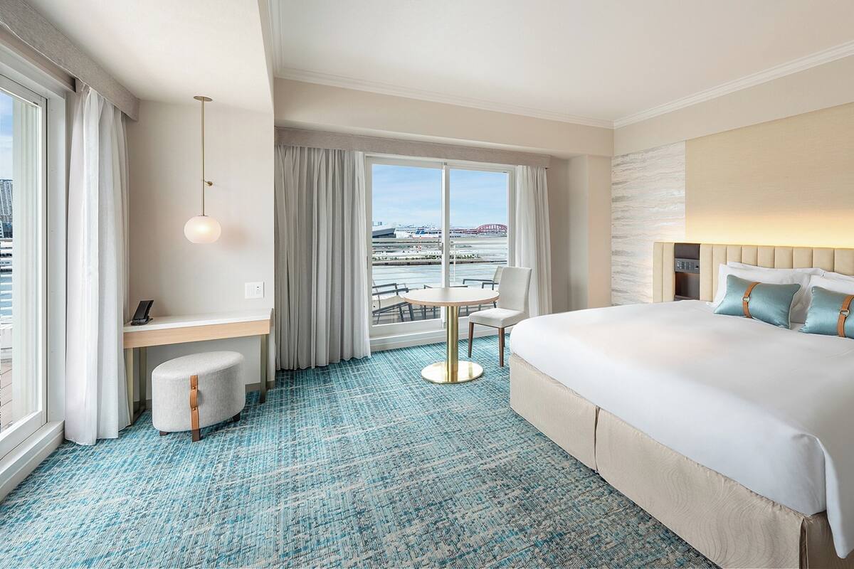 Habitación de hotel con una cama grande, un escritorio con una silla, una mesa redonda y vistas a la ciudad a través de amplias ventanas.