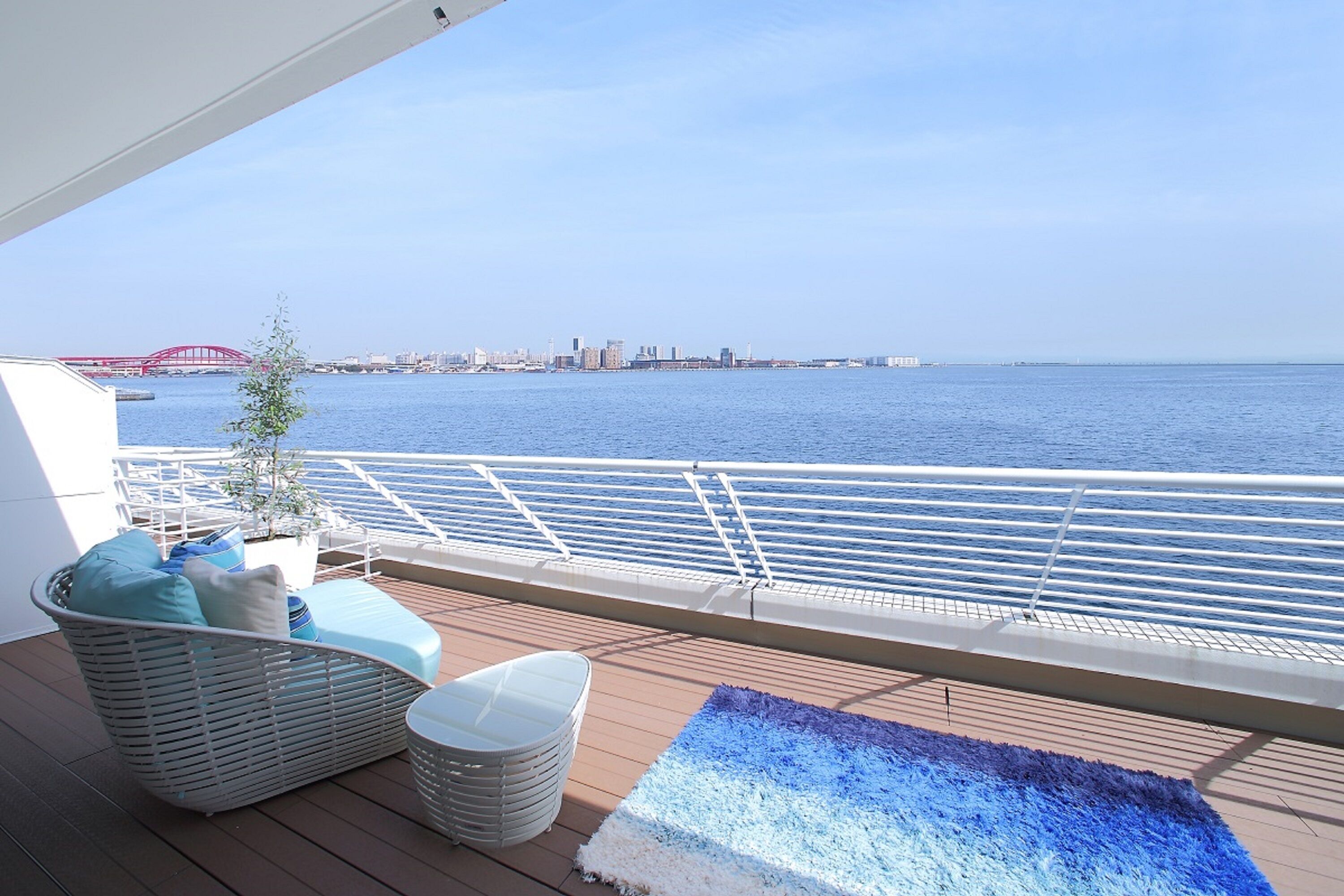 Ein Balkon mit einem Korbstuhl, einem kleinen Tisch und einem blauen Teppich. Im Hintergrund ist eine rote Brücke über dem Wasser zu sehen.