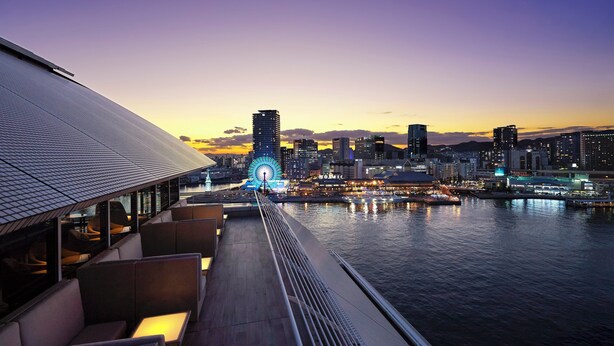 Un restaurante frente al agua con terraza al aire libre y vistas a un horizonte urbano al atardecer.