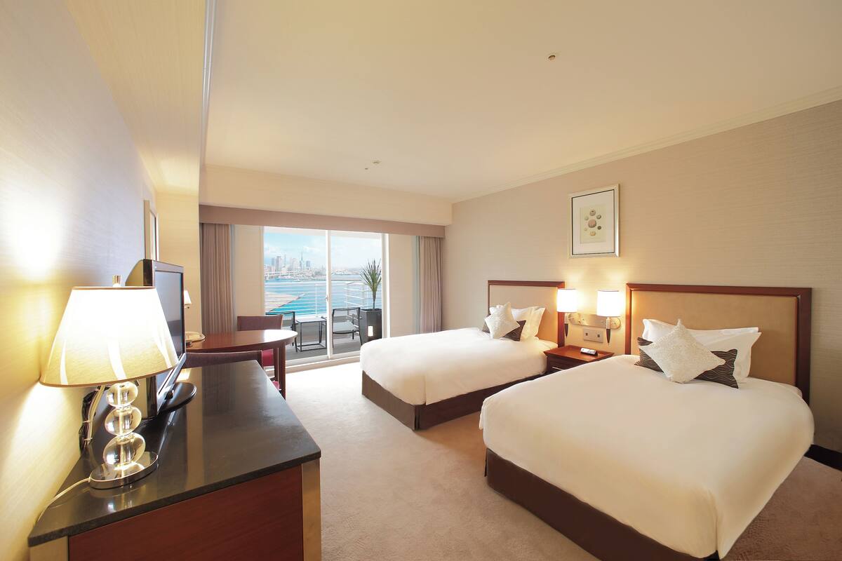 Habitación de hotel con dos camas, un escritorio, una lámpara y vistas a la ciudad.