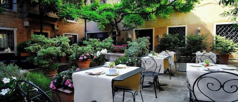 Un patio al aire libre con mesas y sillas, rodeado de plantas en macetas y árboles.