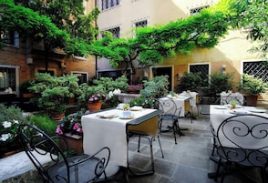 Um terraço ao ar livre com mesas e cadeiras, cercado por plantas em vasos e árvores.