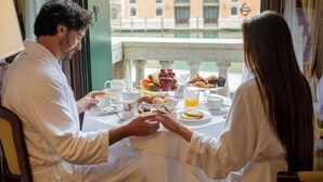 A couple dining at a table with breakfast items, a view of a building, and a window with curtains. - Sina Palazzo Sant'Angelo (Venice)