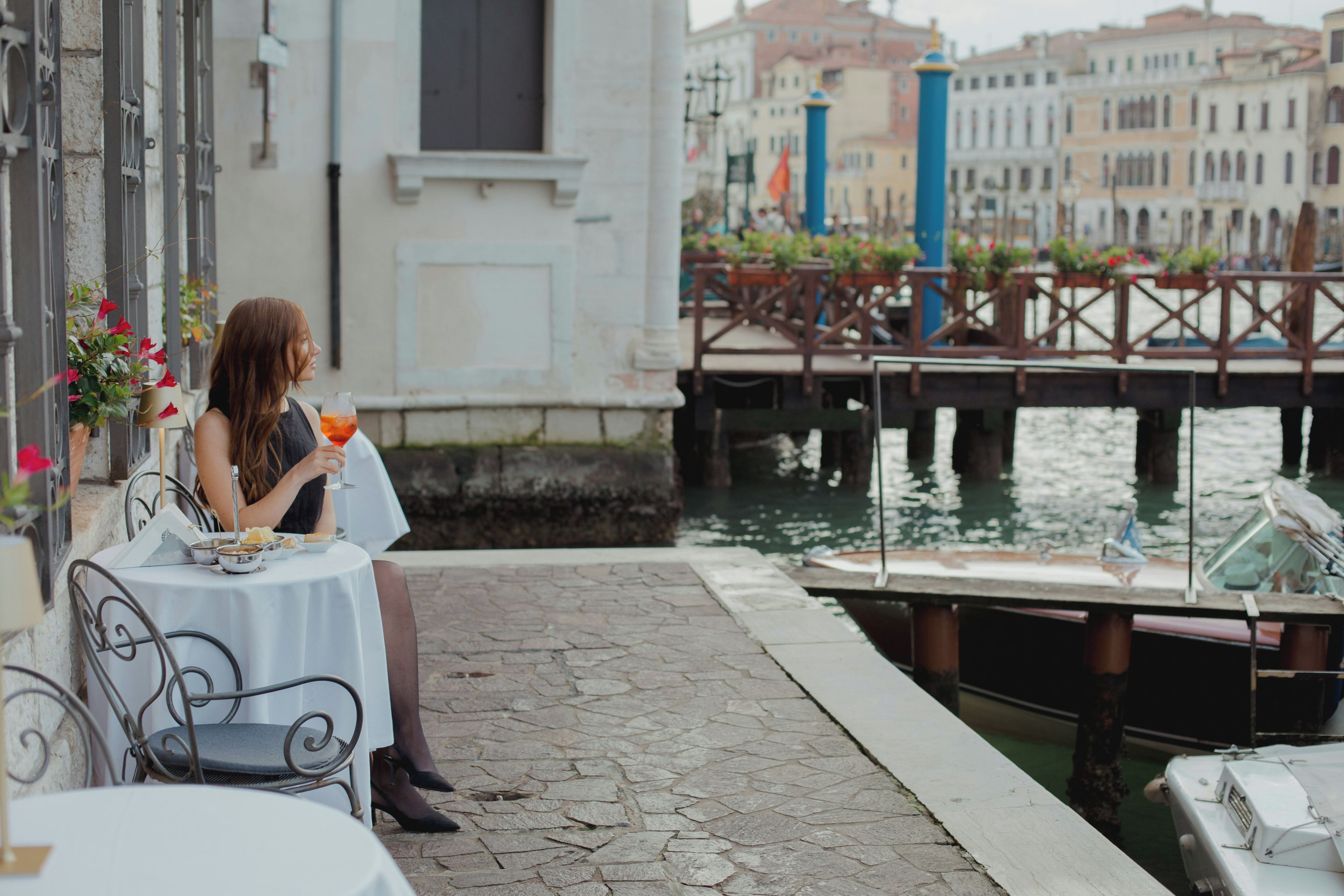 Una mujer sentada a una mesa con una bebida, mirando un canal con barcos y edificios al fondo.